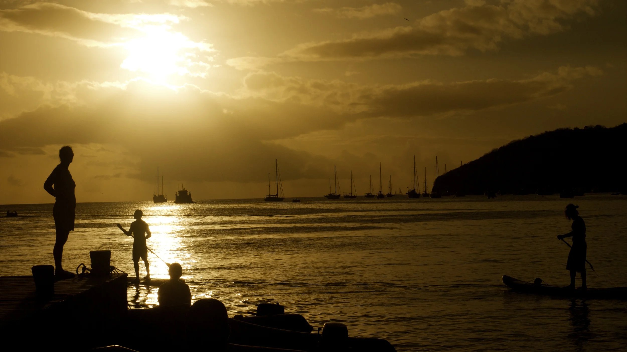 Silhouettes at a Caribbean harbour at sunset — paddleboards, sails, gold water